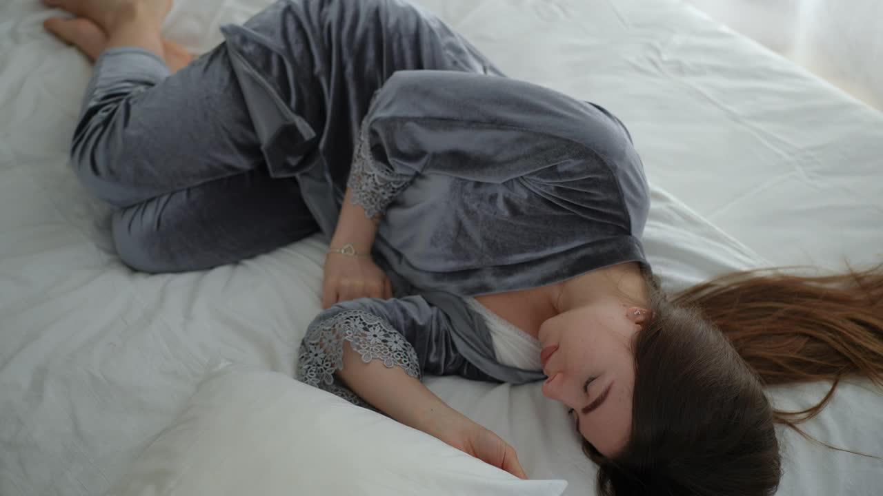 一个穿着漂亮睡衣的女孩躺在床上睡觉。床上有一个枕头。女孩被嘈杂的声音吵醒，用手拿起枕头，盖在头上。视频下载