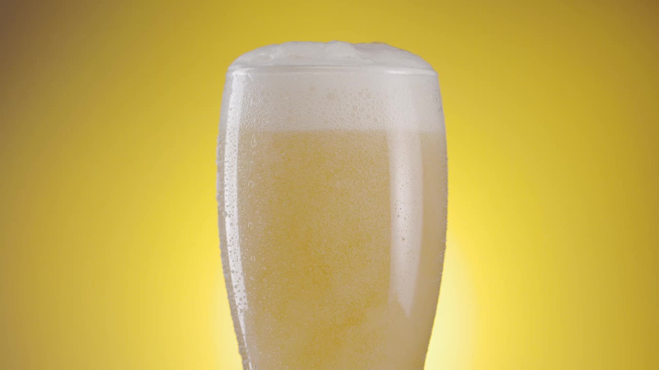 啤酒杯在黄色背景下缓慢旋转，啤酒杯上有凝结，里面有很多气泡和泡沫。一个伟大的啤酒背景为您的项目视频下载