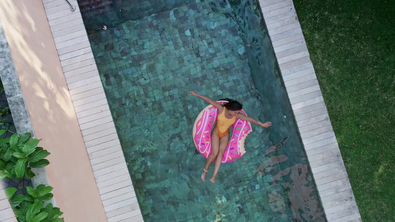 俯视图的年轻女子泳装上粉红色的甜甜圈lilo在游泳池。一名女子在别墅的充气泳池上放松。充气环和床垫。暑假如诗如画。从高处俯瞰…视频下载