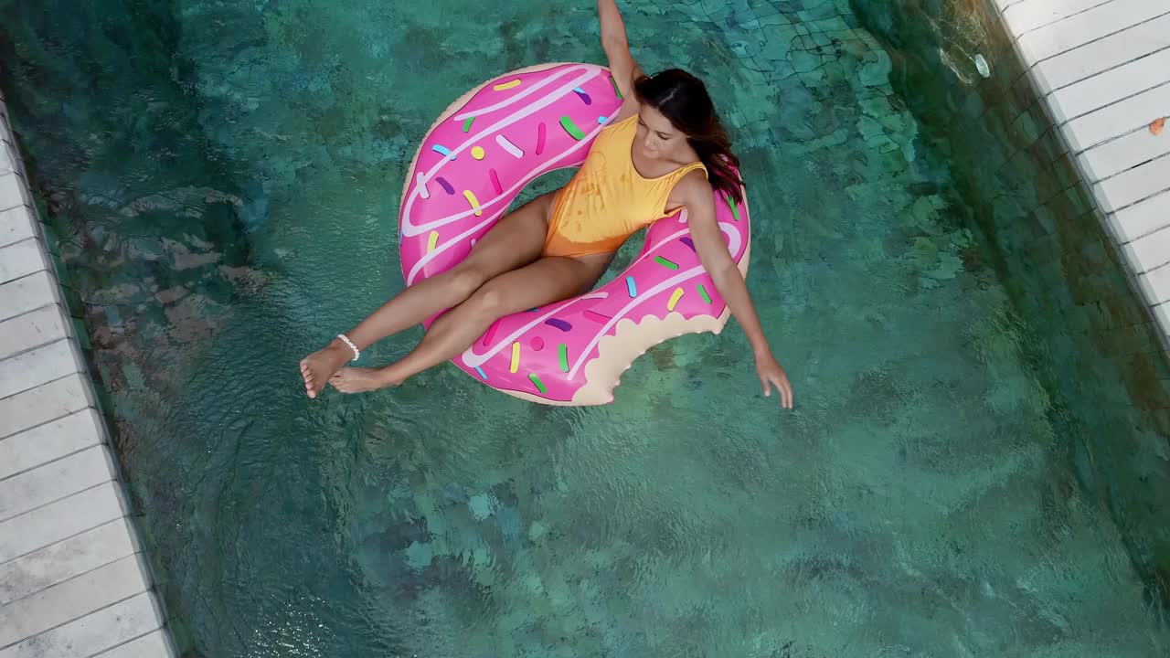 俯视图的年轻女子泳装上粉红色的甜甜圈lilo在游泳池。一名女子在别墅的充气泳池上放松。充气环和床垫。暑假如诗如画。从高处俯瞰…视频下载