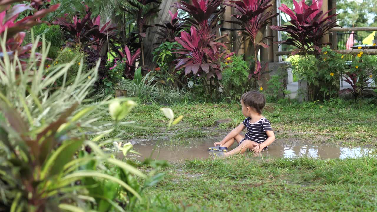 蹒跚学步的男孩坐在院子里的脏水坑里视频下载
