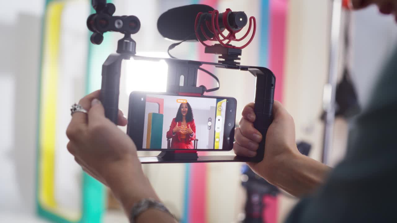 在智能手机上拍摄一位穿着彩色衣服的女电视主持人:时尚的内容创作者与摄影师一起为她的在线频道制作视频的肖像视频下载