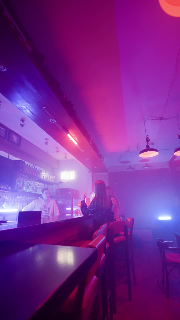 两个女人在夜间酒吧与一名调酒师交谈。紫色和蓝色霓虹灯视频下载