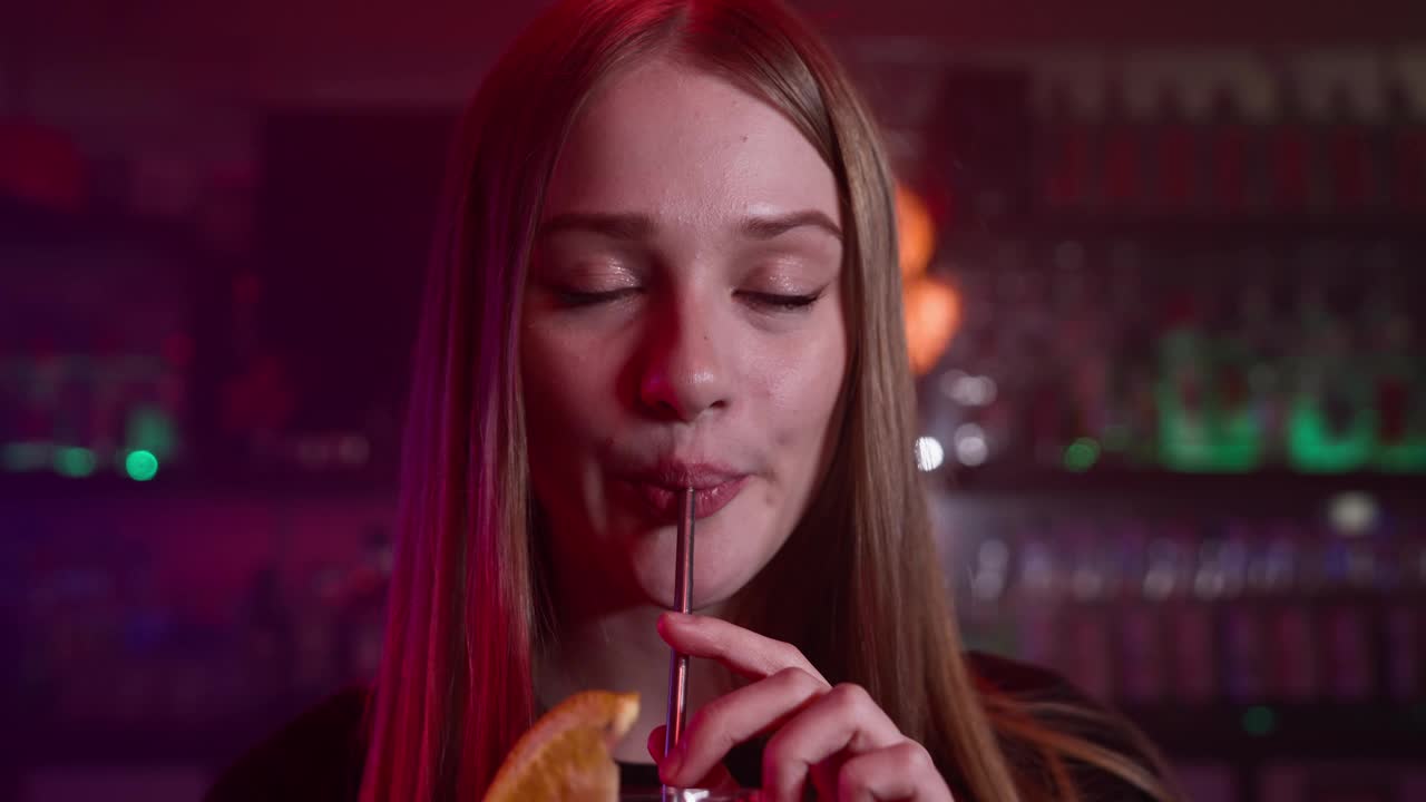 迷人的微笑女孩在霓虹灯下的酒吧喝红色鸡尾酒视频下载