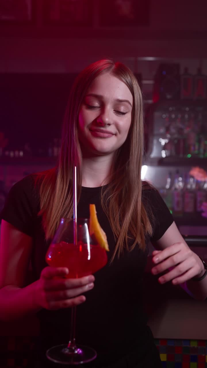 迷人的微笑女孩在霓虹灯下的酒吧喝红色鸡尾酒视频下载