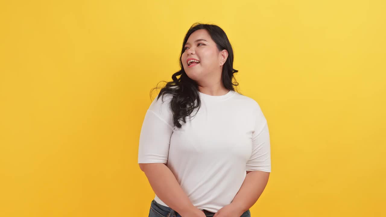 一个胖乎乎的亚洲女人穿着一件白色t恤，在黄色背景上表现出自信和骄傲视频下载