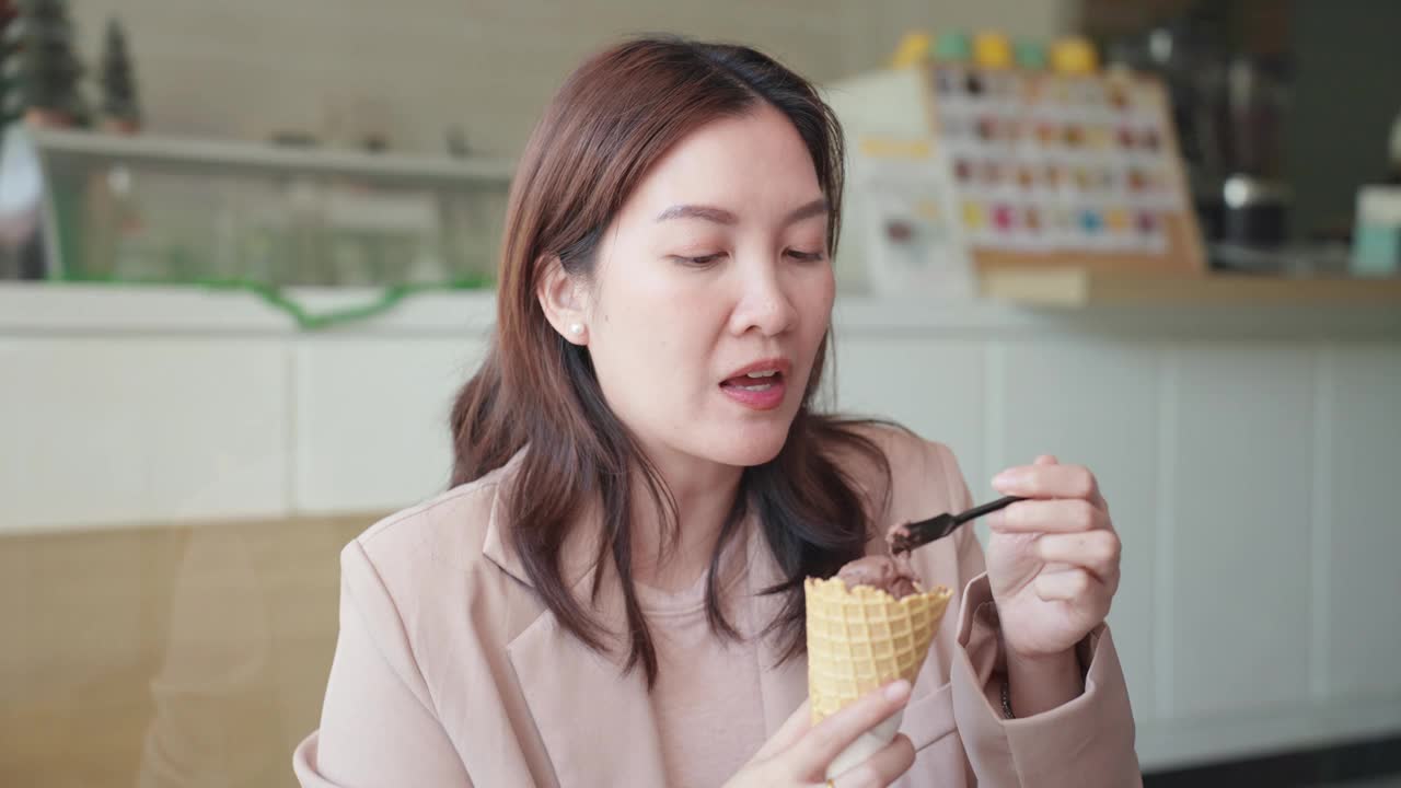 美丽的亚洲女商人为了避免牙痛，用小勺子吃着美味的冰淇淋，但在咖啡馆吃冷甜点时，她仍然感到牙齿敏感和疼痛。牙龈萎缩，牙齿护理。视频下载