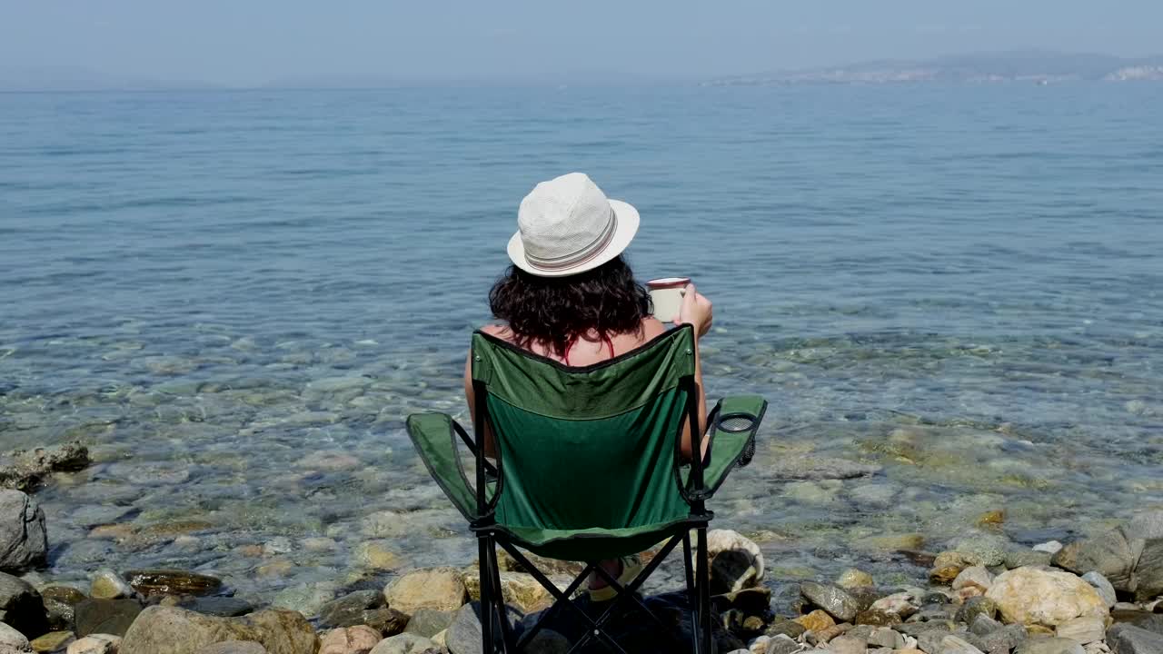 度假的女人坐在沙滩上的露营椅上，享受着一个完美的夏日，喝着咖啡，凝视着大海。节日的概念。视频下载