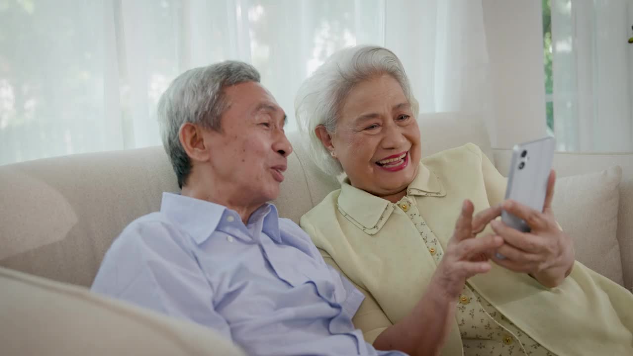 亚洲的老年夫妇喜欢坐在客厅的沙发上玩智能手机视频下载