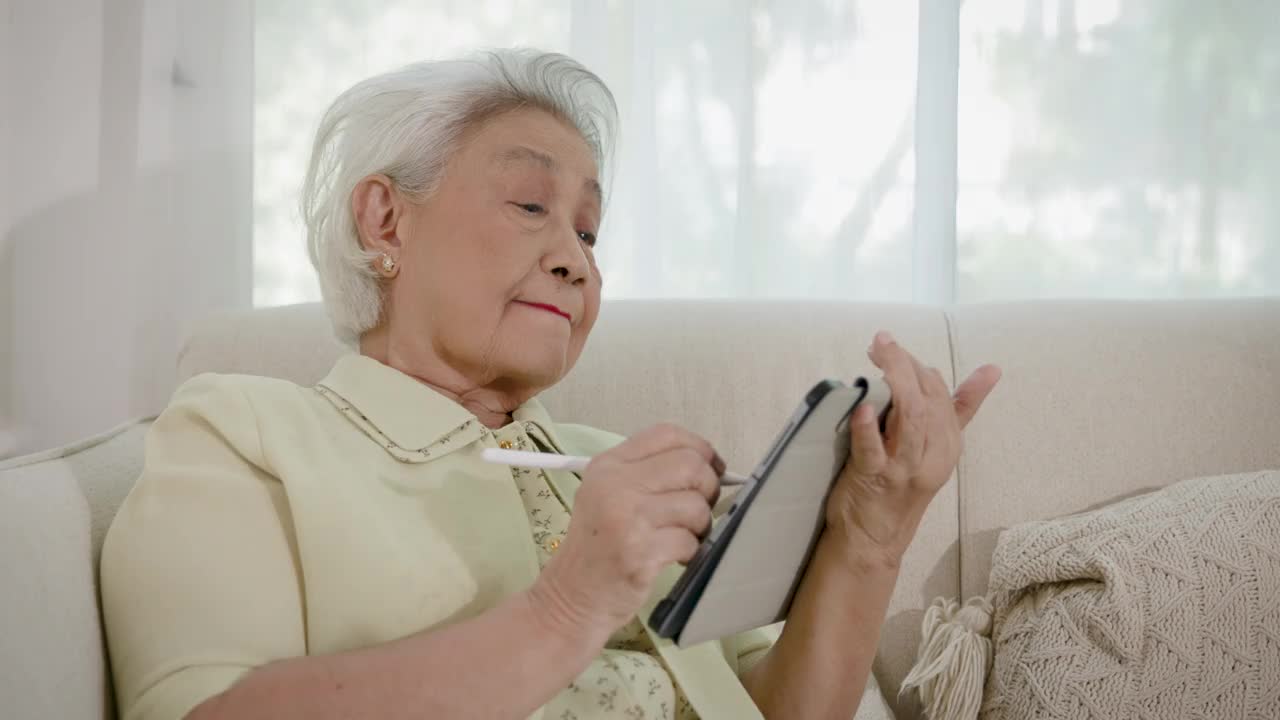 亚洲老年女性喜欢坐在客厅的沙发上玩平板电脑或视频通话视频下载