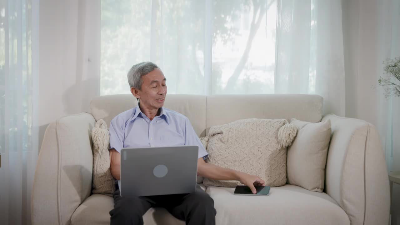 亚洲的老年男性喜欢坐在客厅的沙发上玩笔记本电脑和手机视频下载