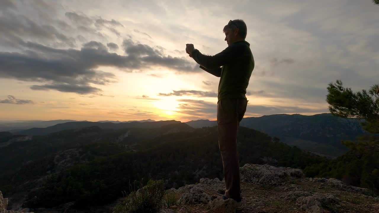 一名40多岁的男子站在山顶上，用智能手机拍摄日落时的山景视频下载