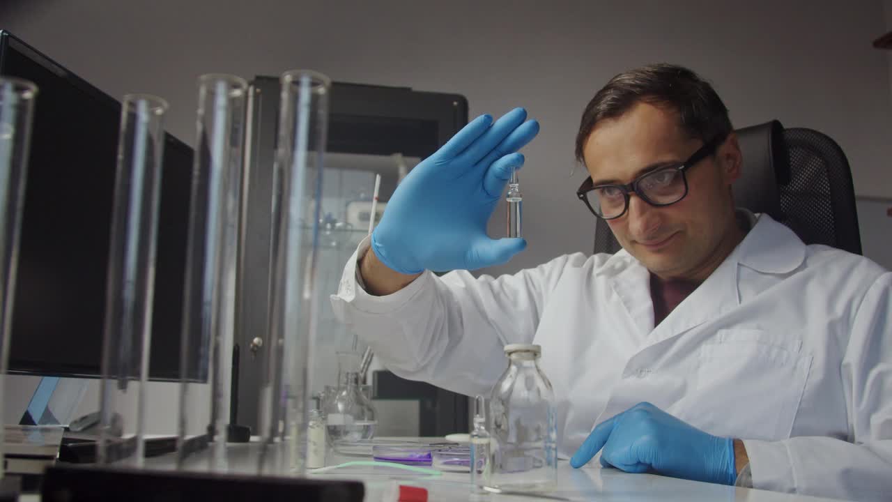 一位年轻的医生穿着实验室工作服，戴着手套，看着一安瓿透明液体的画像。疫苗。保护和保健视频素材