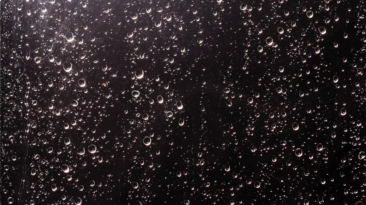 天气预报的屏幕保护程序。雨滴顺着窗玻璃流下来。黑色背景，有各种大小的闪亮球向下流动视频下载
