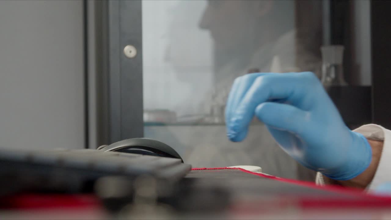 一位男医生在电脑后面工作。一只戴着蓝色防护手套的手使用鼠标的特写。医疗储物柜的侧面反射视频下载