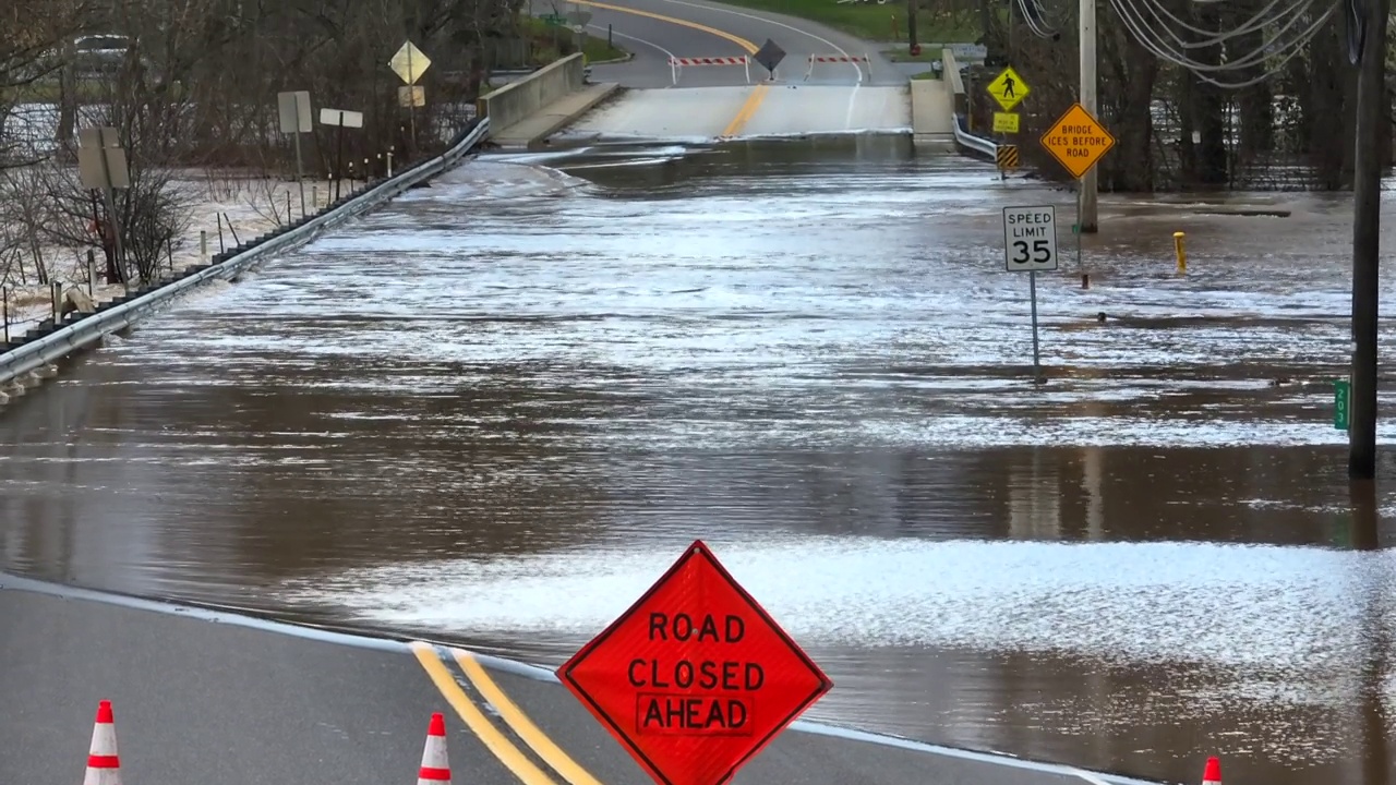 被水淹没的街道前的道路封闭标志和锥形路标。空中倾斜显示河水在桥上泛滥。视频下载