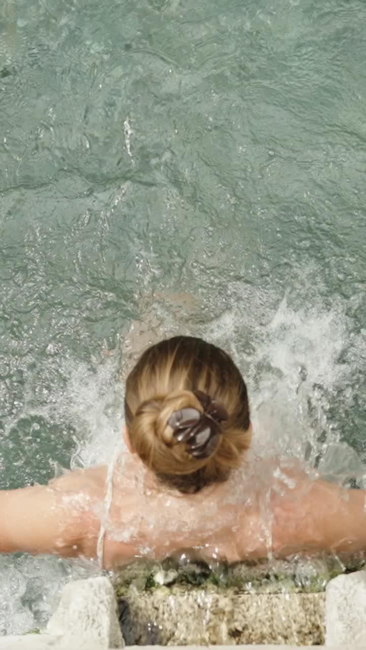 垂直视频。在慢动作中，一位年轻女子站在温泉的水流下，一边享受着这种感觉，一边用手在水中摸索。前视图。视频下载