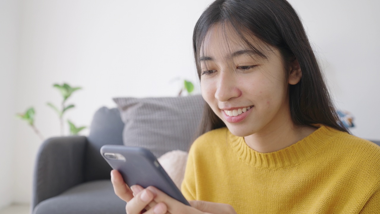 亚洲女孩在家用智能手机快乐地微笑打字视频下载