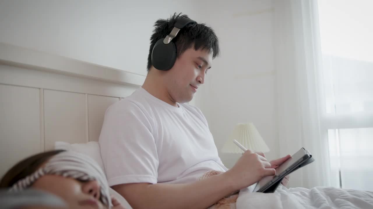 亚洲年轻夫妇躺在床上，女人戴着睡眠面罩睡觉，男人在家里的卧室里用数码平板电脑和耳机听音乐视频下载