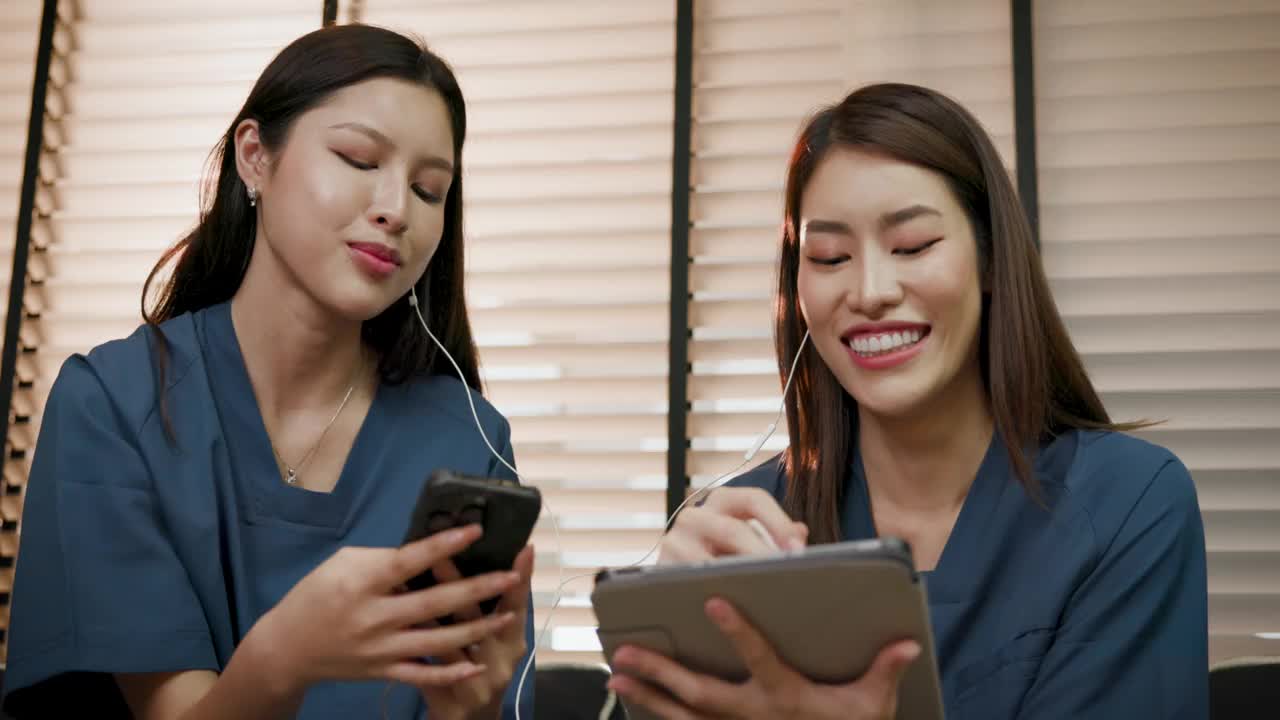 年轻的亚洲医务工作者和同事们下班后坐在诊所或医院的沙发上，用手机和平板电脑休息视频下载