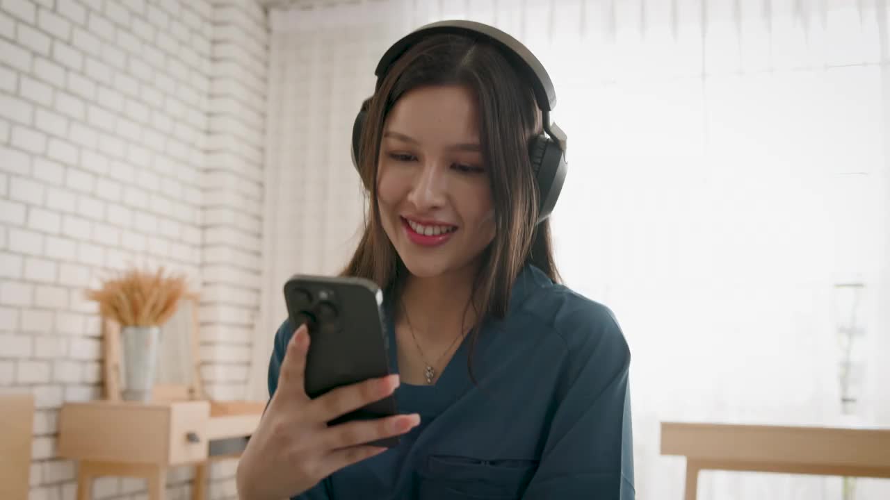 年轻的亚洲医护人员女性穿着手术服，坐在家里的卧室里玩手机，听音乐视频下载