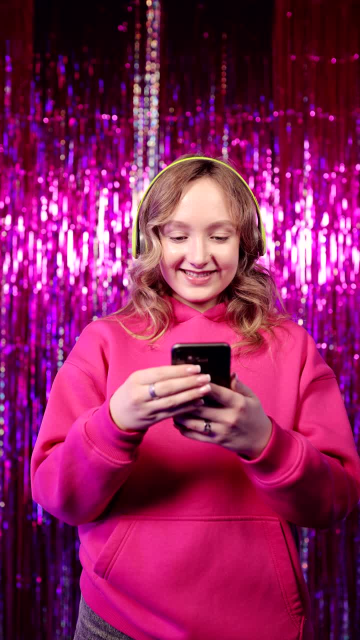 兴奋的年轻女子戴着无线耳机在彩色背景下使用智能手机。在手机里读好消息的快乐女孩。垂直视频。视频下载