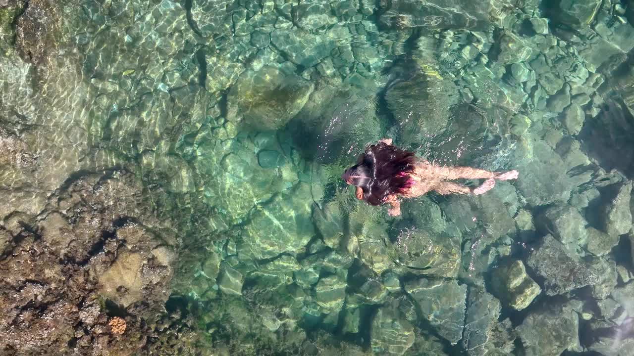 从上面看，一个穿着泳衣的女模特在海边火山岩旁的一个小游泳池里放松。视频下载