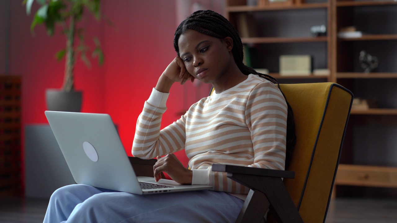 无聊的非洲自由职业者女性坐在家里办公室的扶手椅上，无法集中精力在笔记本电脑上工作视频下载