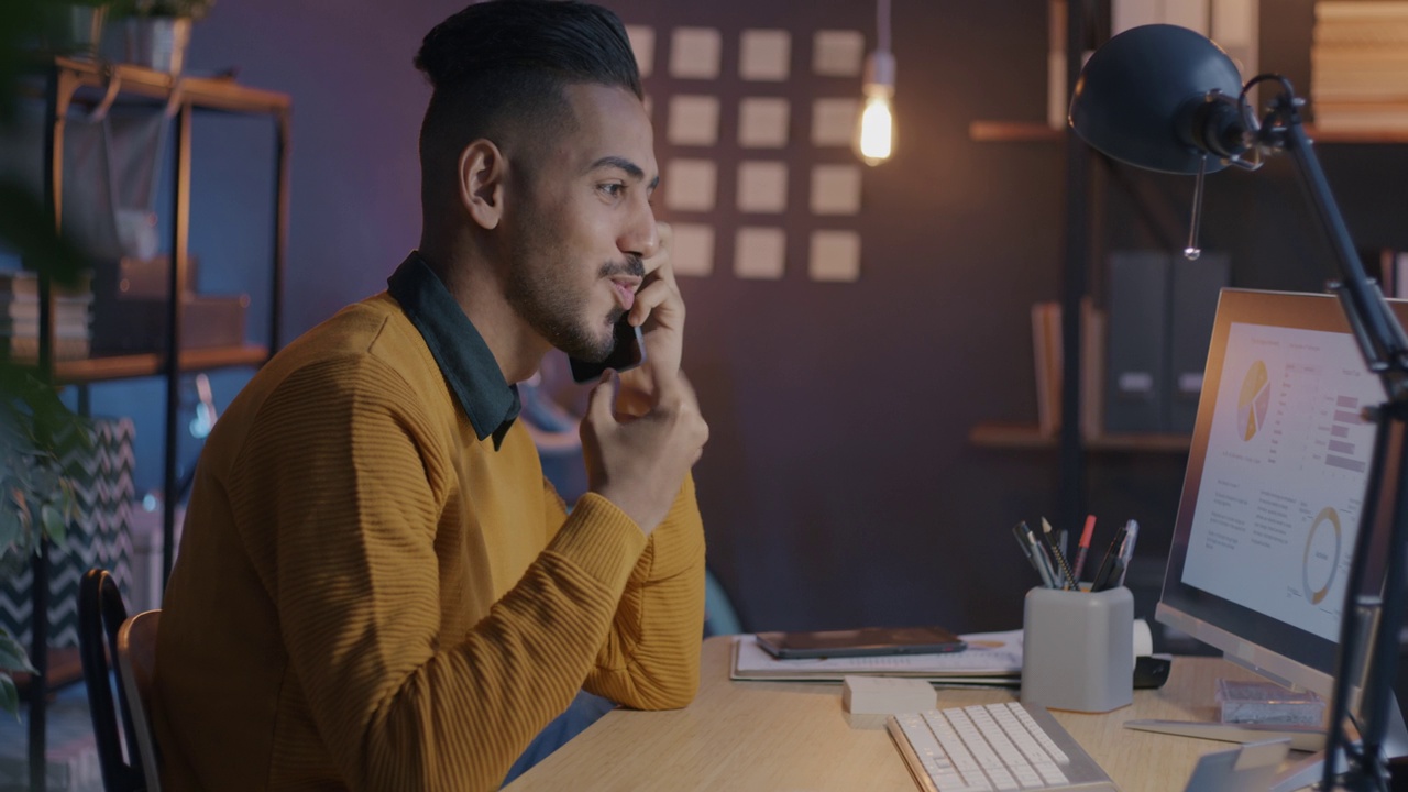 成功的阿拉伯企业家晚上在办公室里打电话，看电脑屏幕上的图表视频素材
