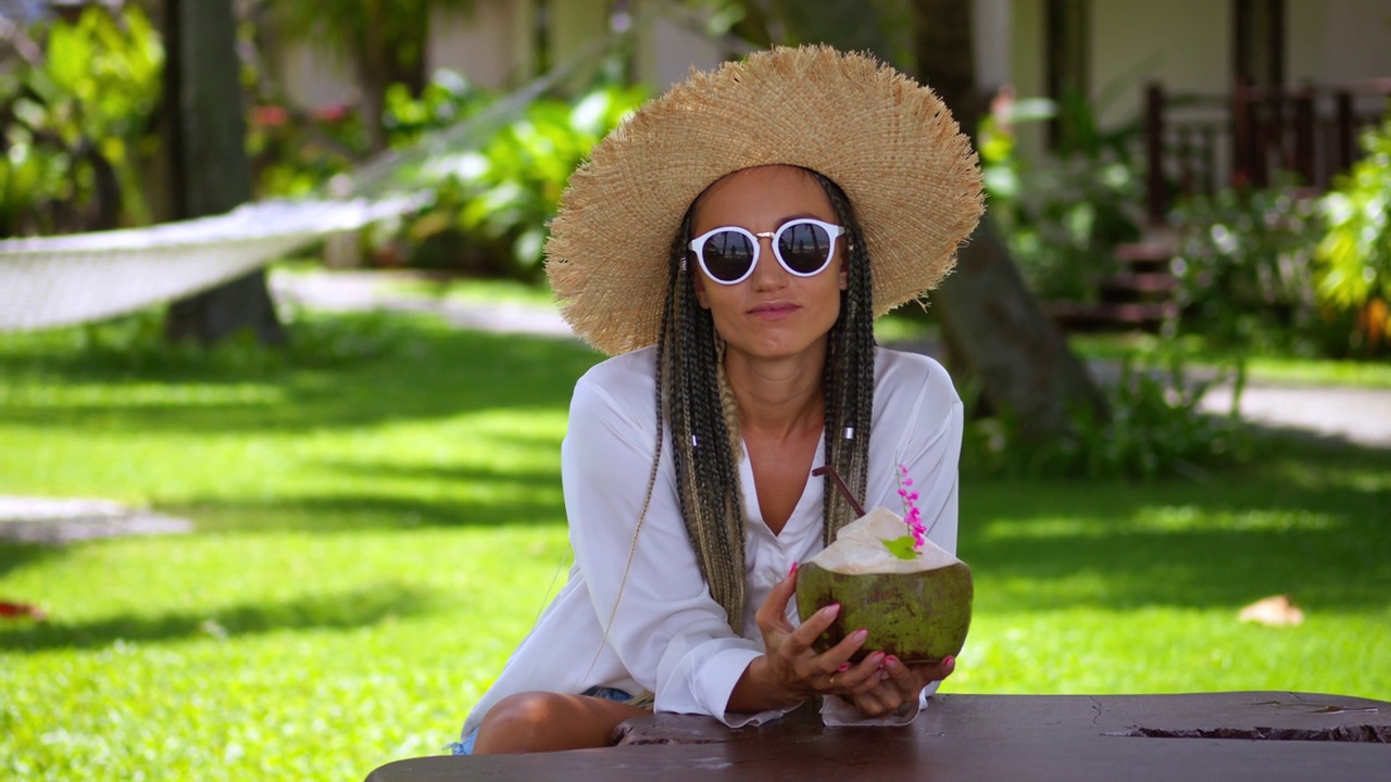 在热带度假胜地，戴着太阳镜和草帽的休闲女游客在海上度假。冰凉的女游客手里拿着新鲜的绿色椰子在户外咖啡馆享受暑假视频下载