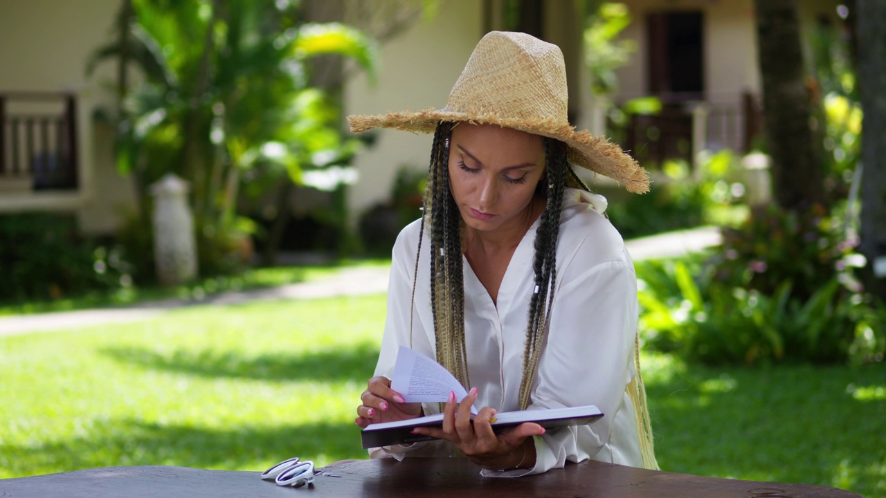 带着草帽的女游客在热带地区的暑假里，在当地户外咖啡馆的桌子上，以棕榈树为背景看书。海风吹动草帽和书页。夏季休闲视频下载
