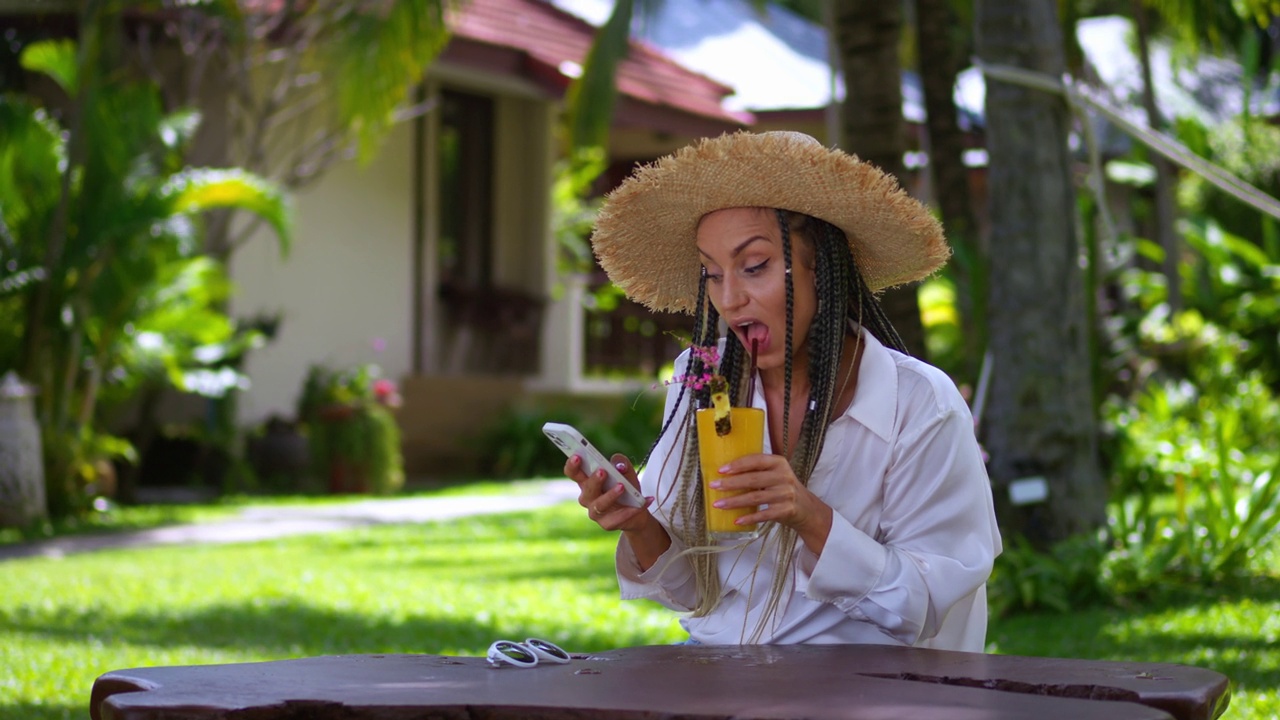 女游客在暑假期间使用手机和喝夏季冷饮。戴草帽的女游客在夏天的户外滚动手机应用程序，阅读新闻，喝香蕉奶昔视频下载