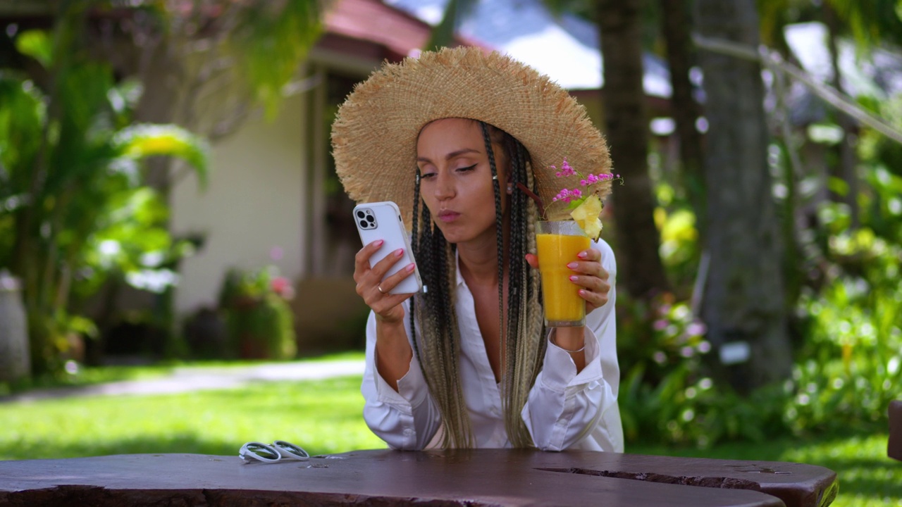 图为女性游客在暑假期间使用智能手机，喝着夏日冷饮。戴草帽的女游客看手机看新闻，夏天在户外喝芒果冷饮视频下载