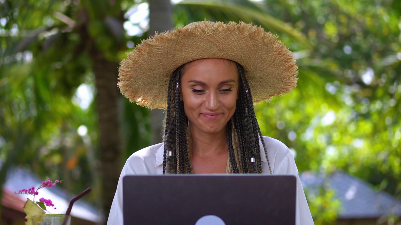 商务女游客在夏季的热带度假胜地用笔记本电脑远程工作。在热带国家旅游的假期里，戴着帽子微笑着用笔记本电脑工作的女商人。远程工作视频下载