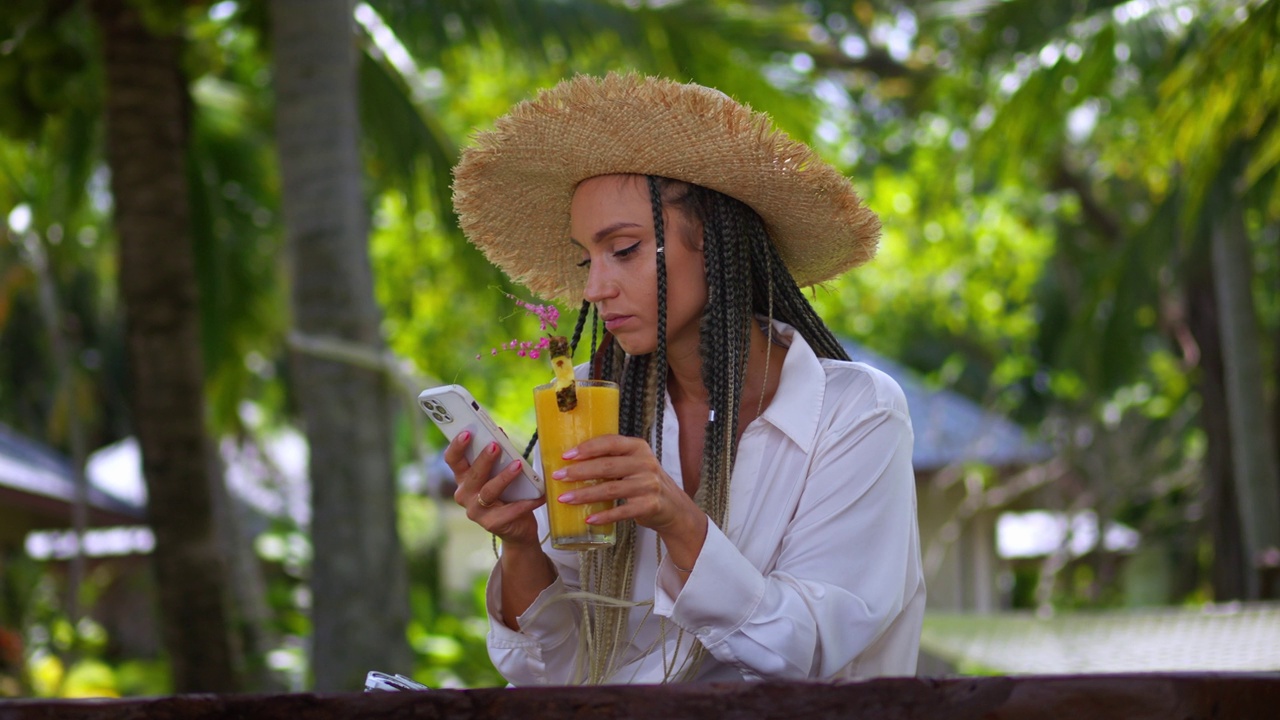 照片中的女性游客在暑假期间使用智能手机，喝着夏日冷饮。戴草帽的女游客翻阅智能手机看新闻，夏天在户外喝香蕉奶昔视频下载