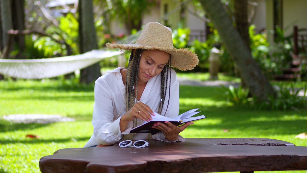 戴着草帽，悠闲的女游客在户外咖啡馆，以棕榈树为背景，在热带度假胜地享受暑假。读书时，微风摇动草帽和书页。暑假视频下载