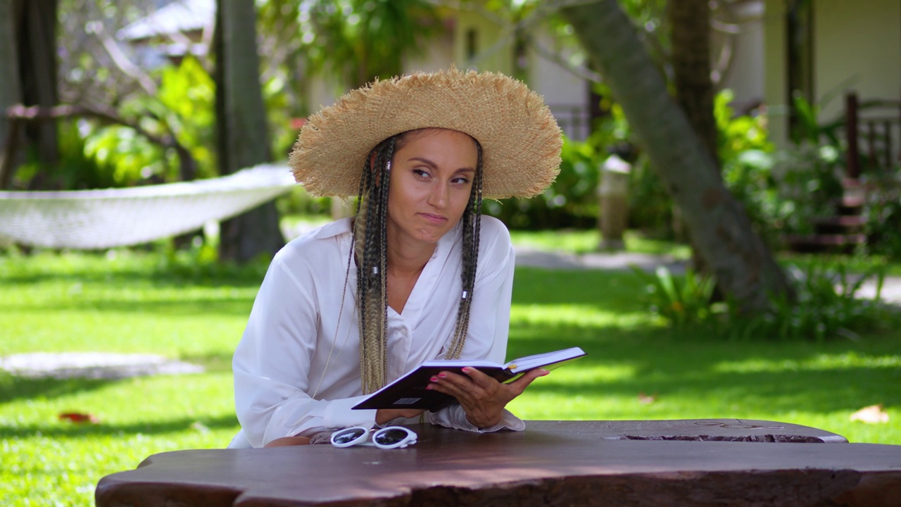 热带度假胜地，戴草帽的女游客在户外咖啡桌旁以棕榈树为背景看书。海风吹动草帽和书页。暑假视频下载