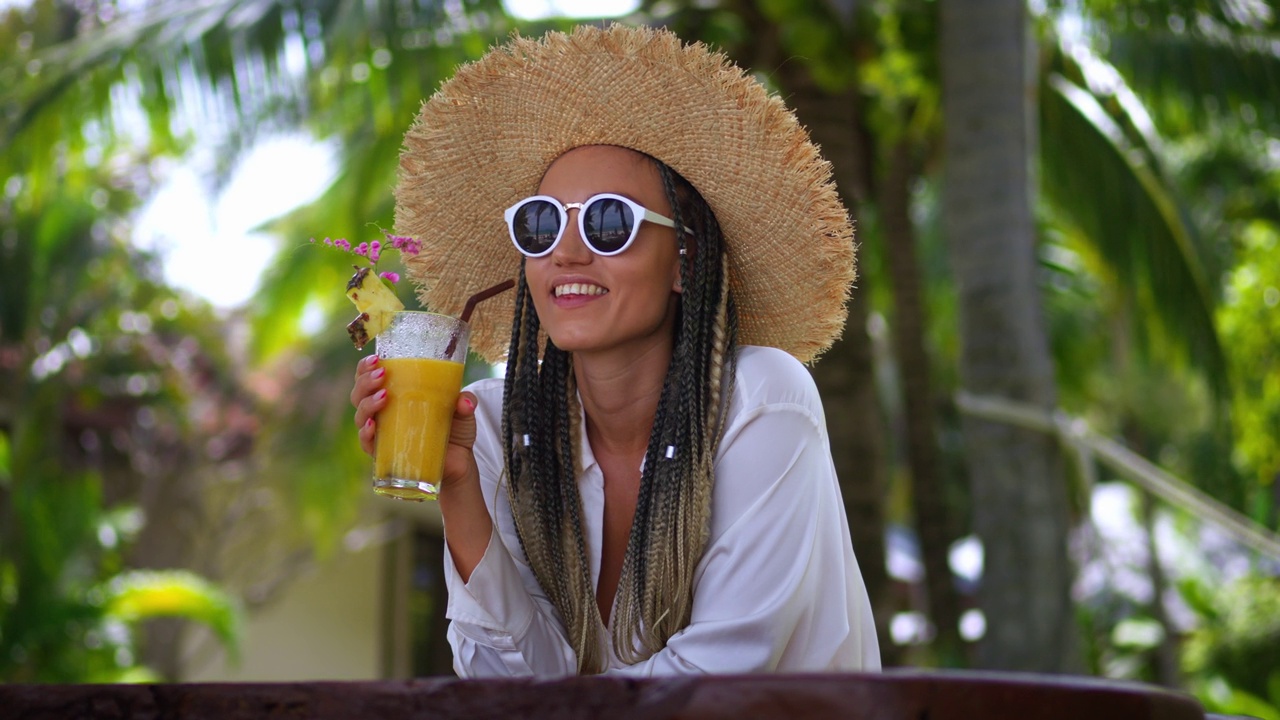 特写:35岁的年轻微笑的女游客，戴着太阳镜和帽子，喝着橙汁，享受着暑假的豪华假期。冷却和休息概念视频下载