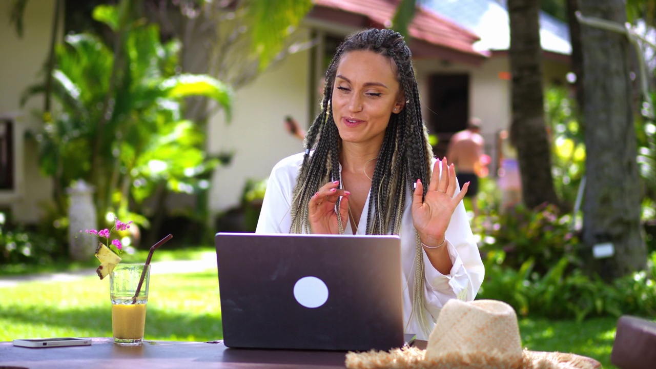 在热带国家度假时，一名微笑的女子通过笔记本电脑上的网络摄像头应用程序进行视频交流。微笑的年轻女子使用笔记本电脑进行在线虚拟交流视频下载