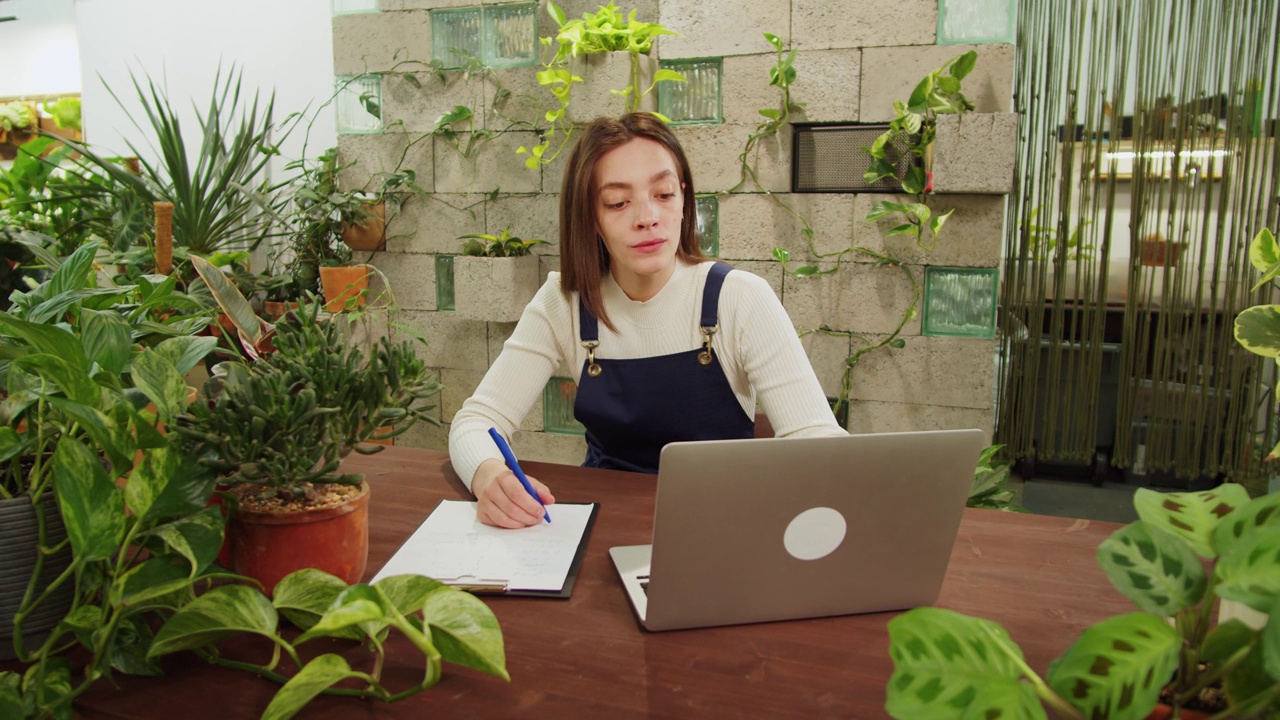 花店女用笔记本电脑视频通话。开虚拟会议的温室工人，写笔记的女植物学家，小企业主。园丁在花店，植物店工作。视频下载
