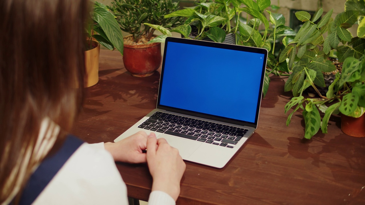 花店女用笔记本电脑视频通话，蓝色度键屏。有虚拟会议的温室工人，女植物学家，小企业主。园丁在花店，植物店工作。视频下载