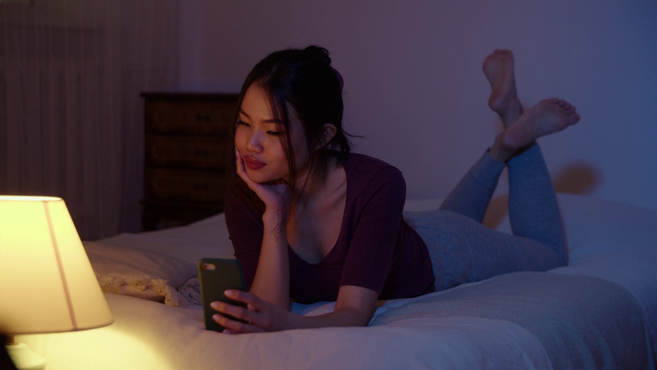 快乐的年轻女子晚上躺在床上玩手机。视频素材