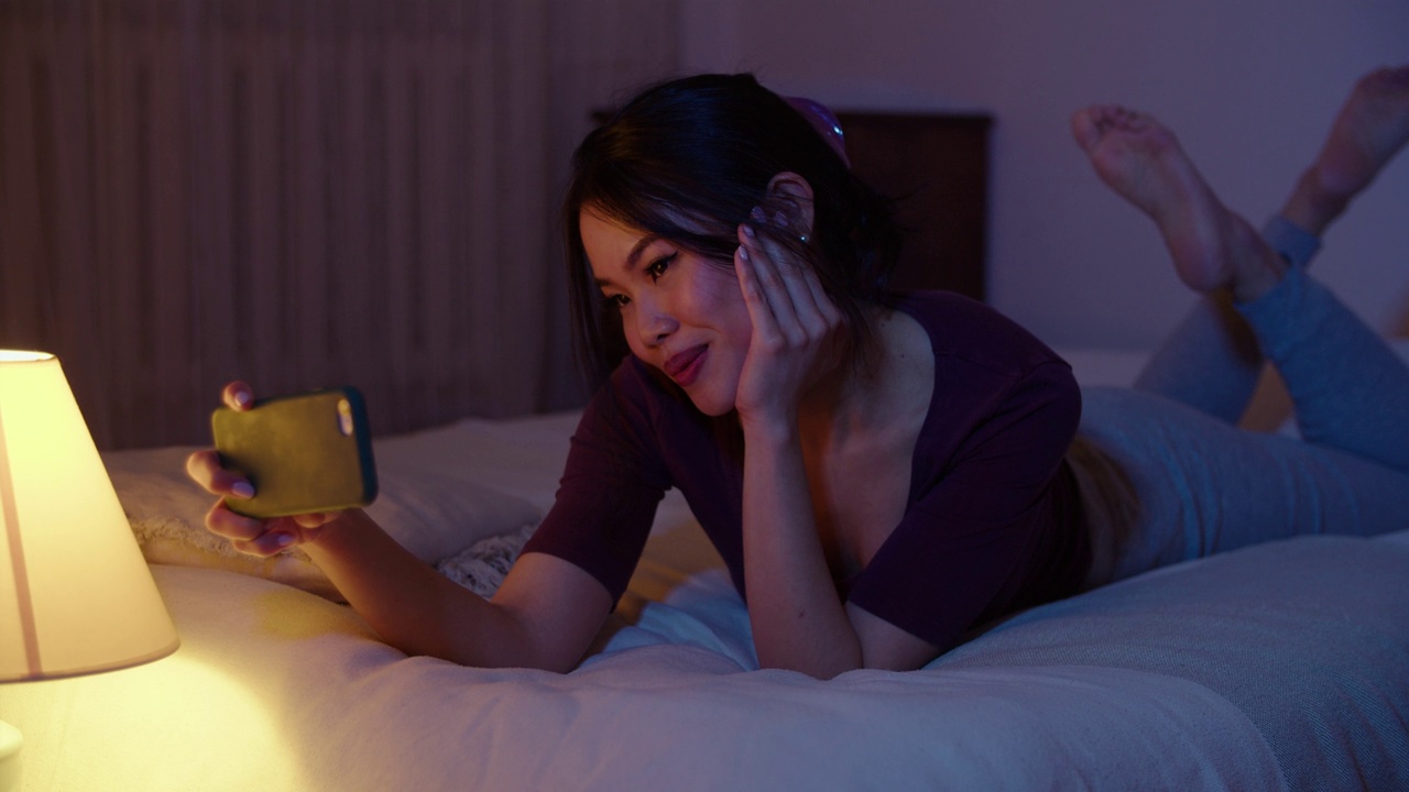 快乐的年轻女子晚上躺在床上玩手机。视频下载