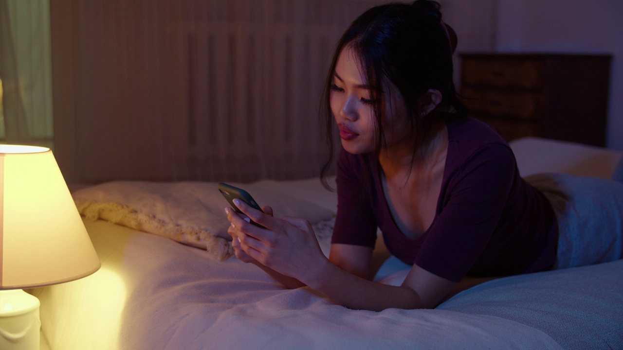 快乐的年轻女子晚上躺在床上玩手机。视频素材