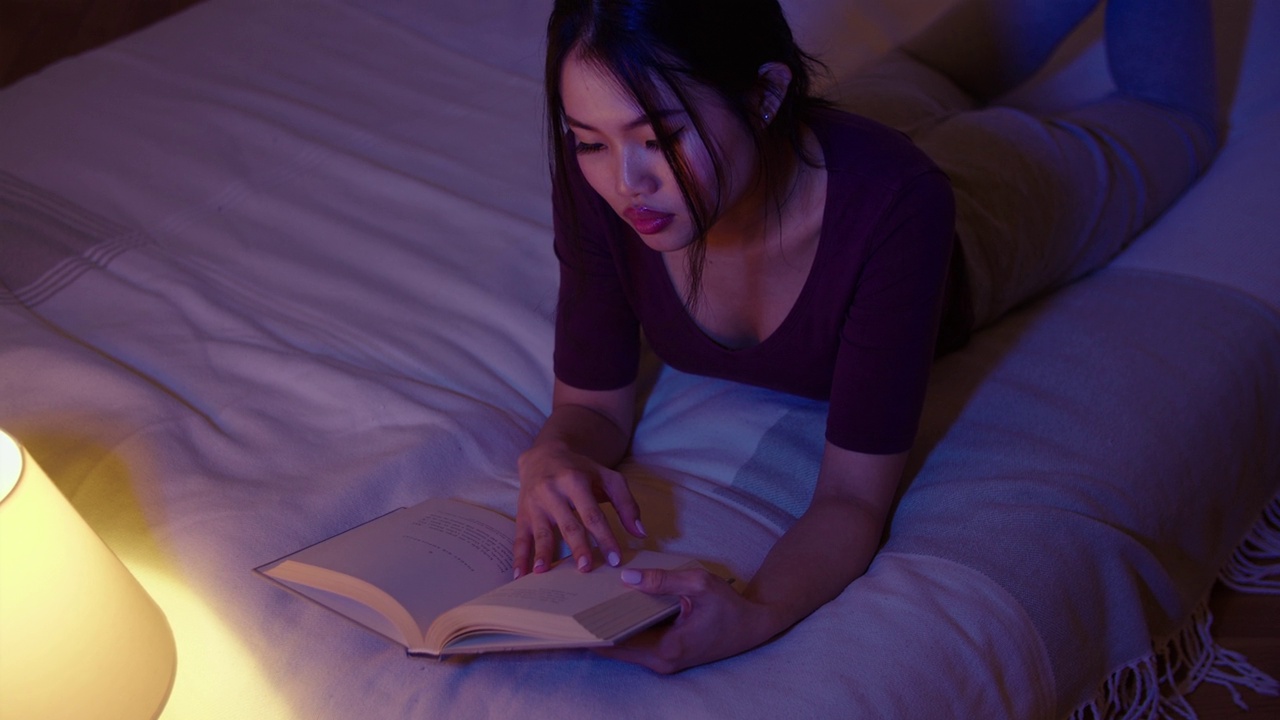 年轻漂亮的女人晚上躺在床上看书。视频下载
