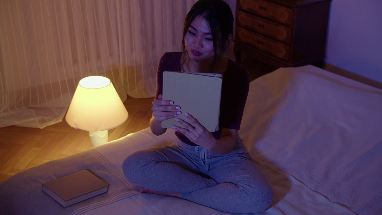 一个微笑的年轻女子躺在床上拿着平板电脑的肖像。视频下载