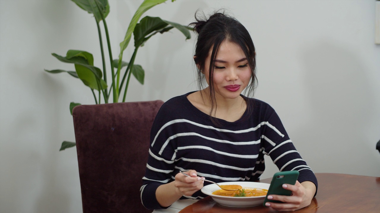 年轻微笑的女人在家里边喝汤边玩手机。视频下载