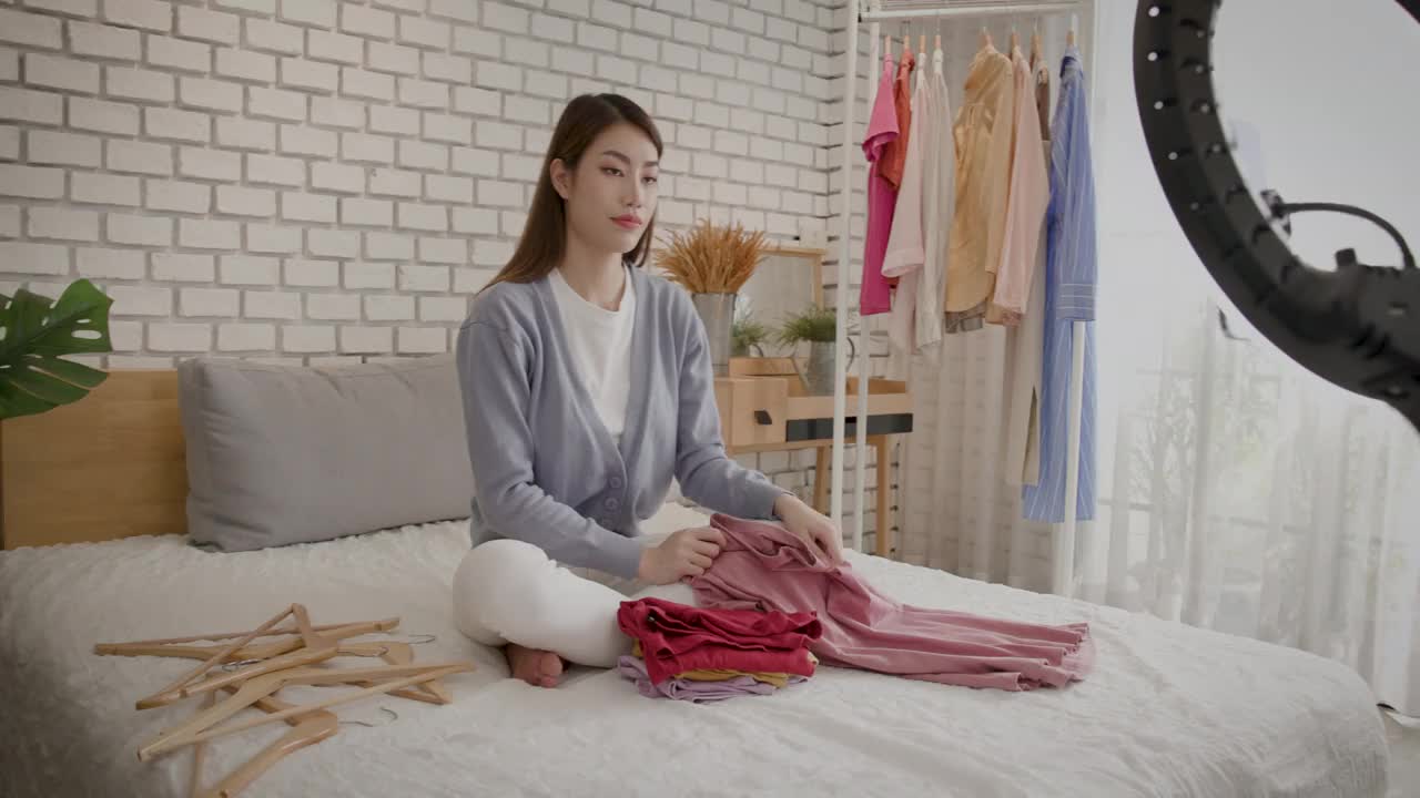 年轻的亚洲女性在家里用手机在社交网络上直播自己卧室里的教程视频，并在网上出售一件衣服视频下载