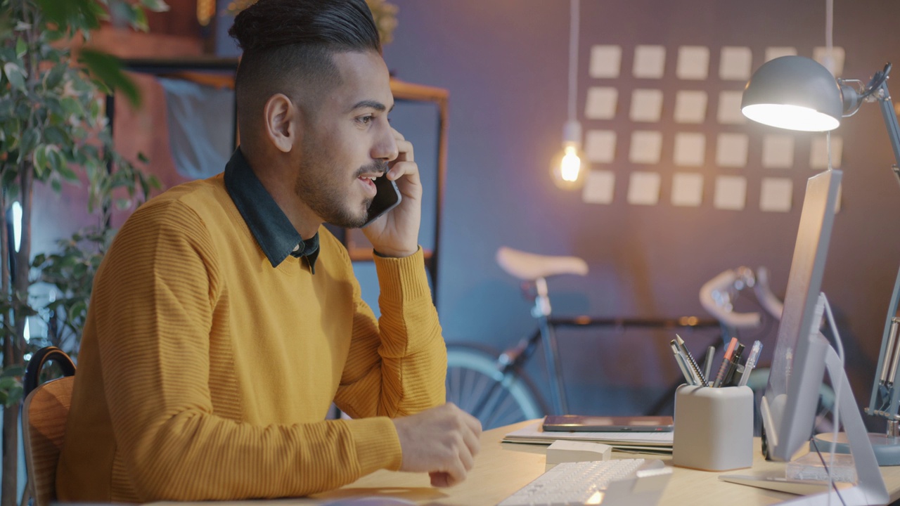 一名中东男子在晚上一边打电话一边看着电脑屏幕讨论办公室的工作视频素材