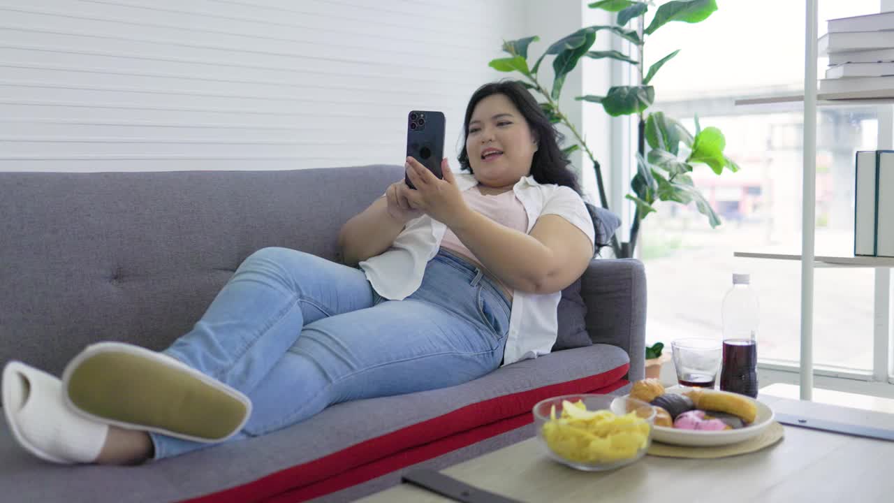快乐的亚洲胖乎乎的女人坐在沙发上微笑，拿着薯片，用智能手机自拍。视频下载